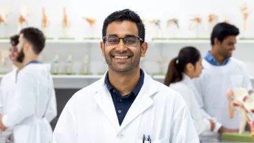 A person in a white lab coat smiling in a lab with colleagues and anatomical models in the background.