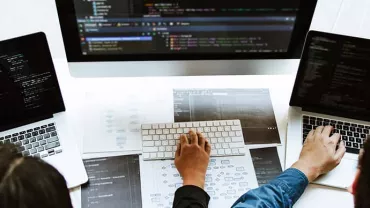 Several people coding on laptops with a large monitor displaying code.