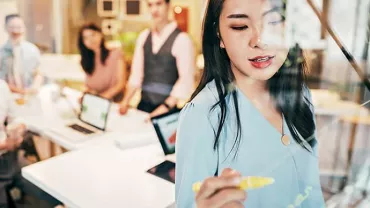 Person in blue shirt writing on glass board during office meeting.