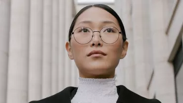 A person wearing formal attire and glasses walks between tall buildings.