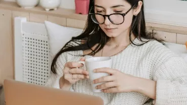 Person on a couch sipping coffee while using a laptop.