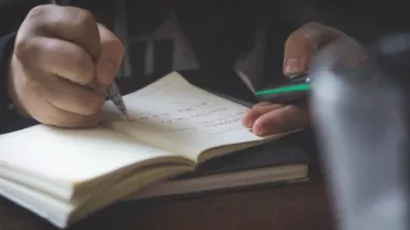 A pair of hands writing in a notebook while holding a phone.