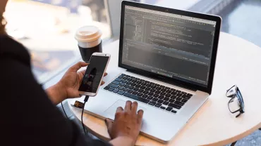 A software developer working on a laptop with a smartphone on a table by the window.