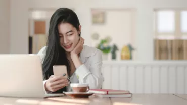 Person smiling at phone with open laptop on table, books, and coffee nearby.