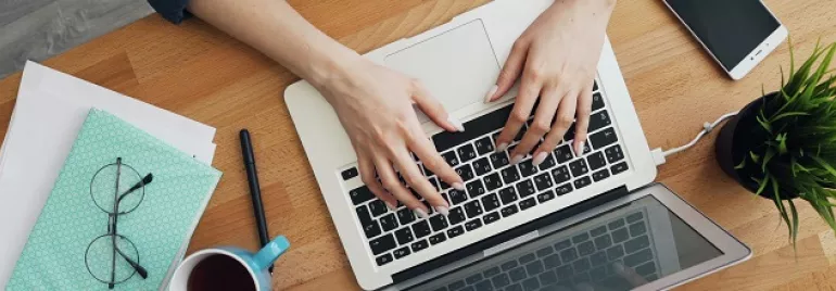 Person typing on a laptop at a wooden desk with glasses, notebook, coffee cup, and plant.